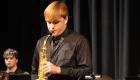Grayson Pinkston performs on saxophone with the Lincoln Trail College Jazz Band, standing on stage under warm lighting in a black dress shirt and tie. He focuses intently on his instrument while a drummer and music stand are visible in the background against a dark curtain backdrop.
