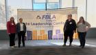 Amie Mayhall, Emelyn Rivera, Jessica Wagner, and Brandy Cochran pose in front of an Illinois FBLA Collegiate State Leadership Conference banner, standing in a bright indoor space with large windows and a cityscape graphic backdrop.