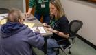 An IECC staff member sits with an older adult at a table, reviewing paperwork together and offering guidance, while another staff member stands nearby assisting.