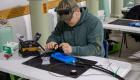 A Broadband Telecom student sits at a workstation carefully splicing fiber optic cables, using specialized tools and equipment spread across a table during hands-on training.