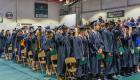 Graduates stand together during Lincoln Trail College’s Commencement Ceremony in the LTC Sports Center as they move their tassels to mark the completion of their degrees and certificates. Family members and guests watch from the bleachers in the background.