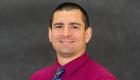A portrait of Ryan Hawkins. He has short, black hair and is wearing a magenta shirt with a blue tie.