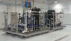 Industrial process training skid featuring two large black tanks, interconnected blue and white piping, valves, gauges and a control panel, mounted on a metal platform with access stairs.