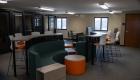 Collaborative workspace at The 618, featuring green lounge seating, orange ottomans, high-top tables and white chairs with orange cushions. Glass-walled meeting rooms line one side of the space.