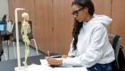 Students sits at lab table looking at table top skeleton modle while writing in her notebook.